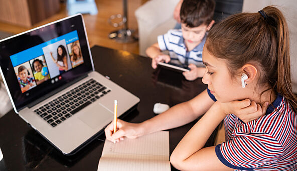 A photo of a student participating in remote learning on her computer with a sibling next to her on a tablet.